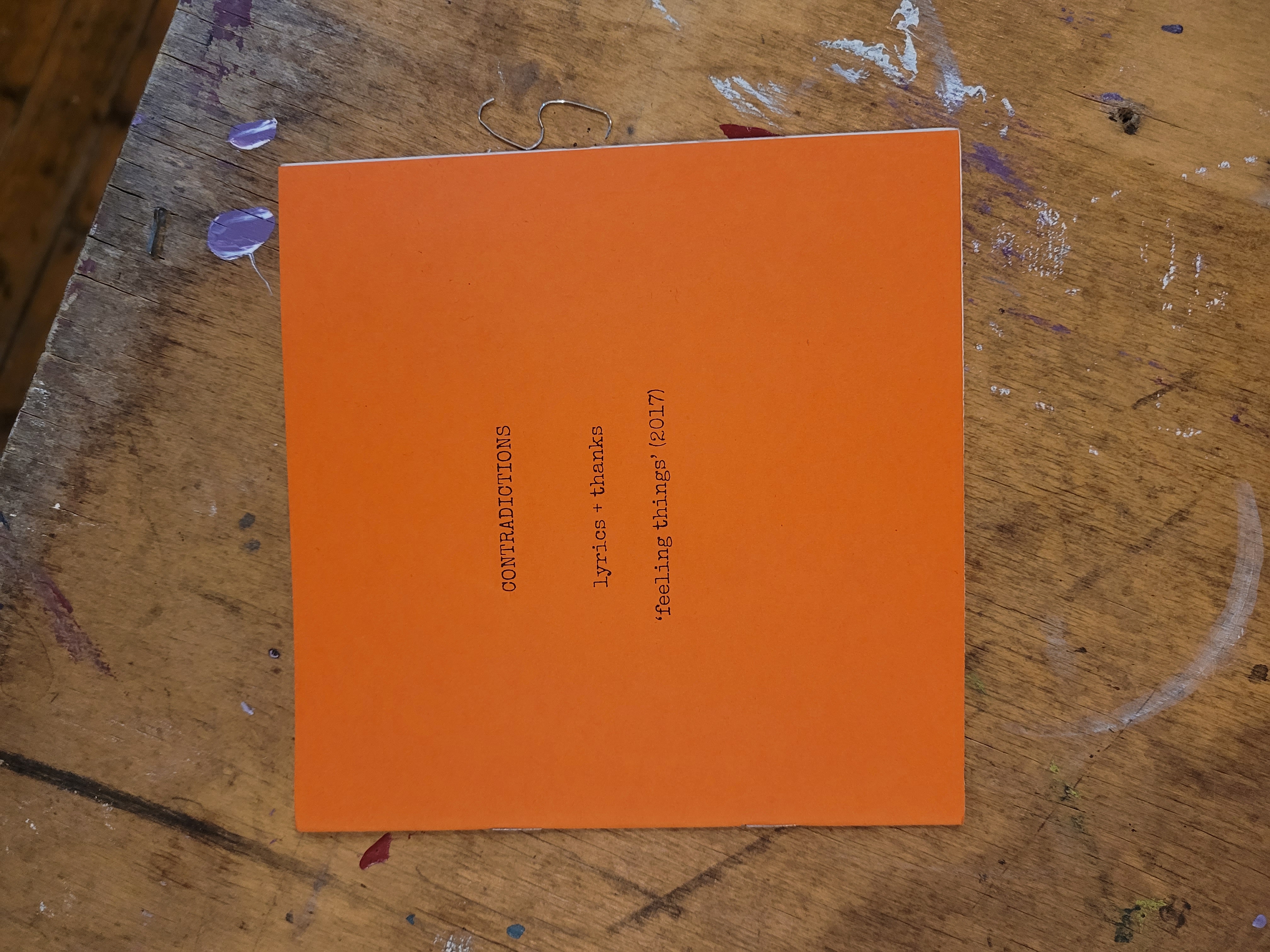 square bright orange zine laying on table. Typewriter text reads "CONTRADICTIONS" lyrics + thanks, 'feeling things' 2017.