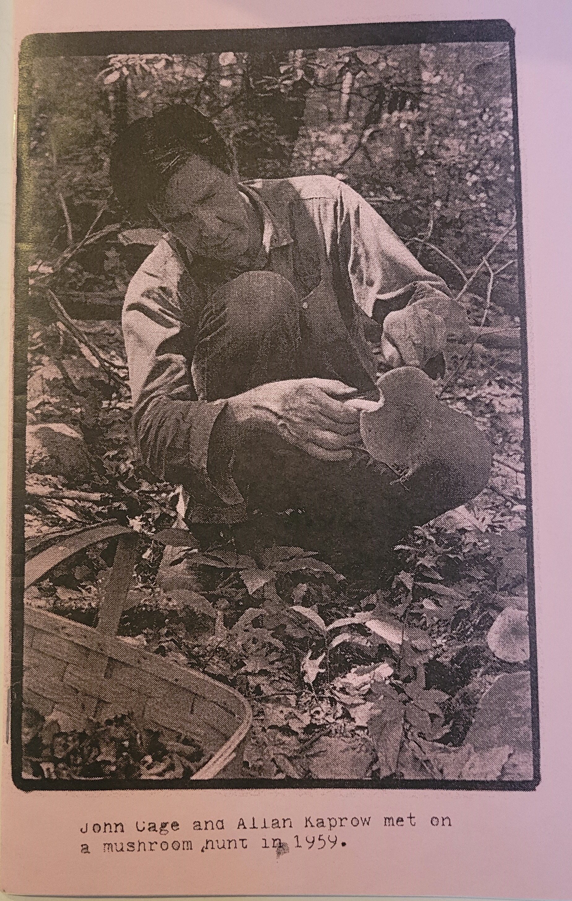 Black and white photo on pink paper, man bending down in forest looking at gills of mushroom, wicker harvesting basket in bottom corner of image