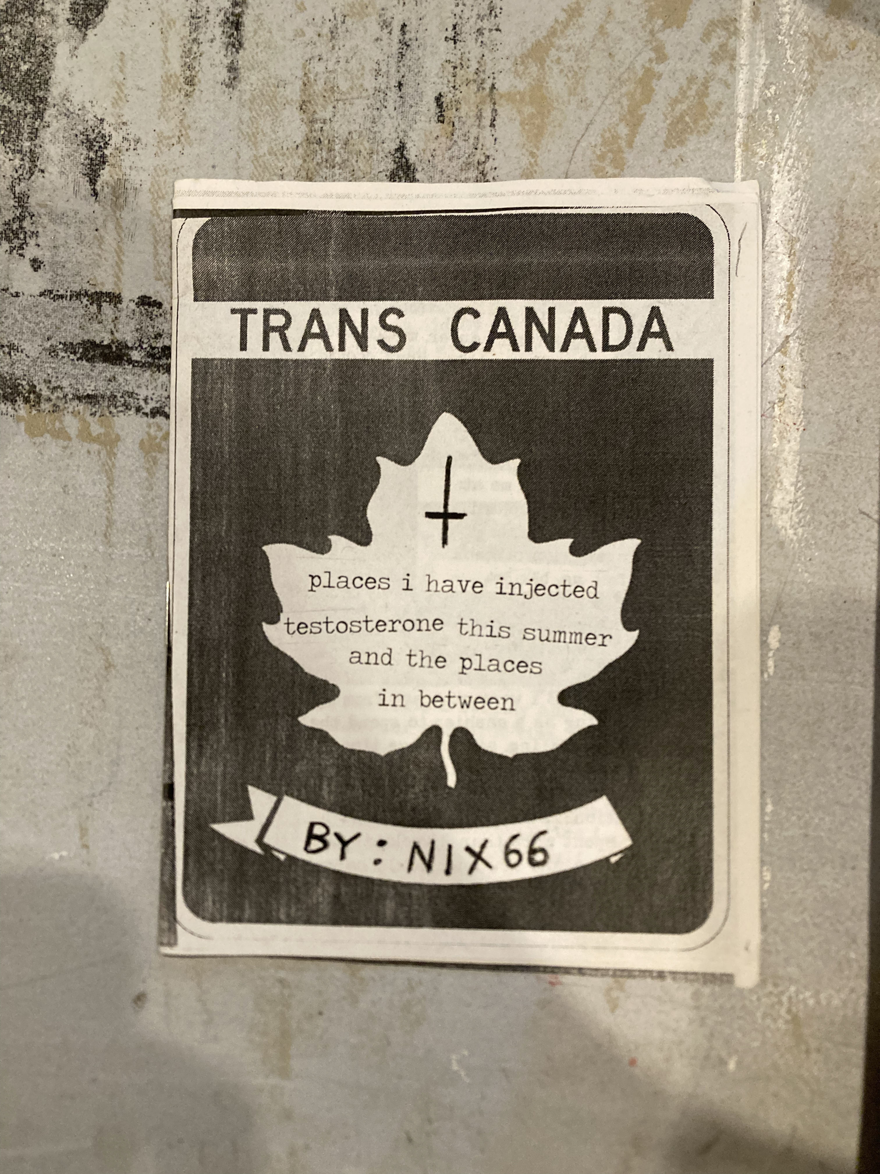 Black and white photo of a road sign with a white maple leaf in the middle. The leaf has an inverted cross drawn on it.