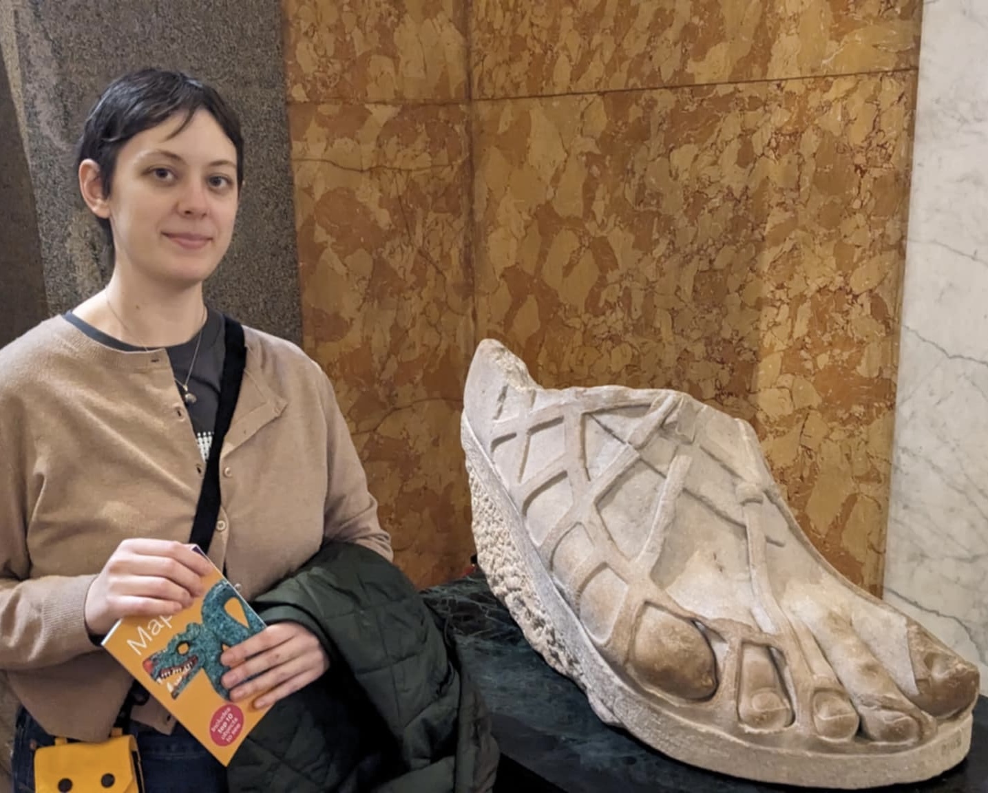 Photograph of a person with short brown hair and a light brown sweater, holding a yellow book and a black jacket, standing beside a sculpture of a foot